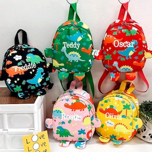 May include: Five colorful dinosaur-shaped backpacks with personalized names: Freddie, Teddy, Oscar, Rosie, and Olivia. The backpacks are made of fabric and have a variety of dinosaur designs.