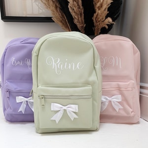 May include: Three personalised backpacks with white bows. The backpacks are green, purple and pink. The names "Raine", "Esme-Rae", and "LM" are embroidered on the front of the backpacks.