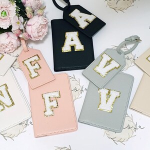 May include: Set of six luggage tags and passport holders with different letters of the alphabet. The tags are made of a soft, textured material and feature a white letter with gold glitter accents. The passport holders are made of a similar material and feature a white letter with gold glitter accents.