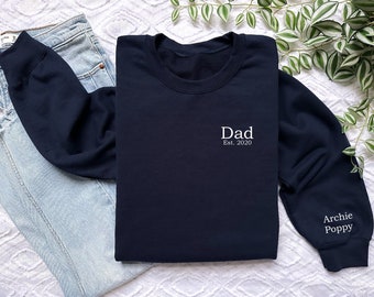 Może przedstawiać: Granatowa bluza z napisem "Dad Est. 2020" na piersi. Na prawym rękawie widnieje biały napis "Archie Poppy". Bluza jest złożona obok pary niebieskich dżinsów.