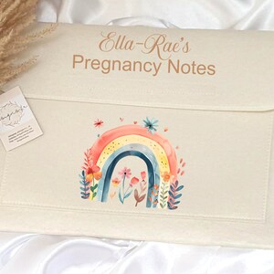 May include: A white felt folder with a rainbow and flowers design. The folder has the text "Ella-Rae's Pregnancy Notes" printed on the front.