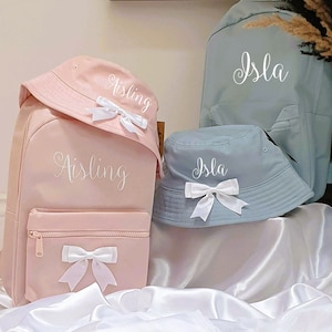 May include: Two pink and blue backpacks with matching bucket hats. The backpacks and hats are personalised with the names "Aisling" and "Isla" in white cursive lettering.