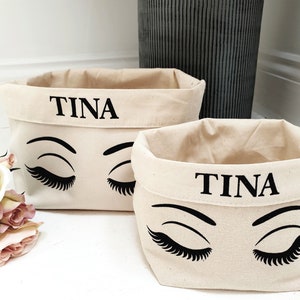 May include: Two beige canvas storage baskets with black eyelashes and the name "TINA" printed on the front.