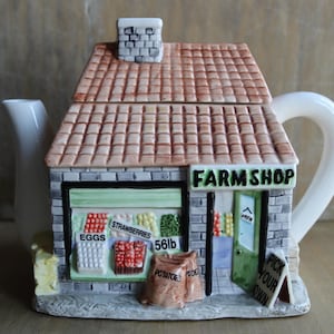 Może przedstawiać: Ceramiczny czajniczek w kształcie wiejskiego sklepu z brązowym dachem z dachówek i kominem. Z przodu widnieje napis FARM SHOP, EGGS, STRAWBERRIES, 56lb, POTATOES oraz PICK YOUR OWN. Posiada białą wylewkę i uchwyt.