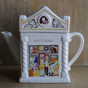Puede incluir: Una tetera de cerámica blanca con forma de edificio y tapa en forma de tejado. El frente presenta una colorida ilustración del interior de una tienda de antigüedades con el texto "ANTIQUES" arriba y "an antique teapot" en la base.