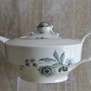 Op de afbeelding: Een crèmekleurige theepot van keramiek met een verfijnde botanische illustratie van groene en grijze bloemen en bladeren. De theepot heeft een bijpassend deksel met een zilverkleurige knop, een gebogen handvat en een taps toelopende tuit.