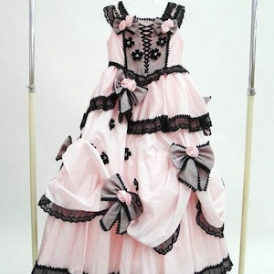 Könnte beinhalten: Ein rosa Satin-Kleid mit schwarzer Spitzenborte und Schleifen. Das Kleid hat ein tailliertes Oberteil mit Schnürung im Rücken und einen vollen Rock. Das Kleid ist mit schwarzen und rosa Blumen und Schleifen verziert.