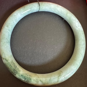Könnte beinhalten: Ein glatter, kreisförmiger Armreif aus hellgrünem Jade mit natürlichen, gesprenkelten weißen und dunkelgrünen Einschlüssen im gesamten Stein. Die polierte Oberfläche reflektiert das Licht und unterstreicht die Transparenz des Materials.