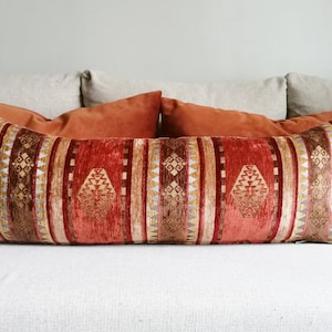 May include: A long, rectangular throw pillow with a red, brown, and gold geometric pattern. The pillow is on a white couch with two smaller orange pillows behind it.