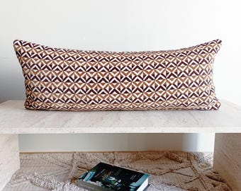 May include: A long, rectangular decorative pillow with a geometric pattern in shades of brown, tan, and white. The pillow is placed on a light-colored surface, showcasing its design and shape. The pillow's dimensions are approximately 30 inches long.