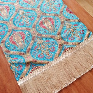 May include: A turquoise table runner with a paisley pattern and gold accents. The runner has a fringe border.