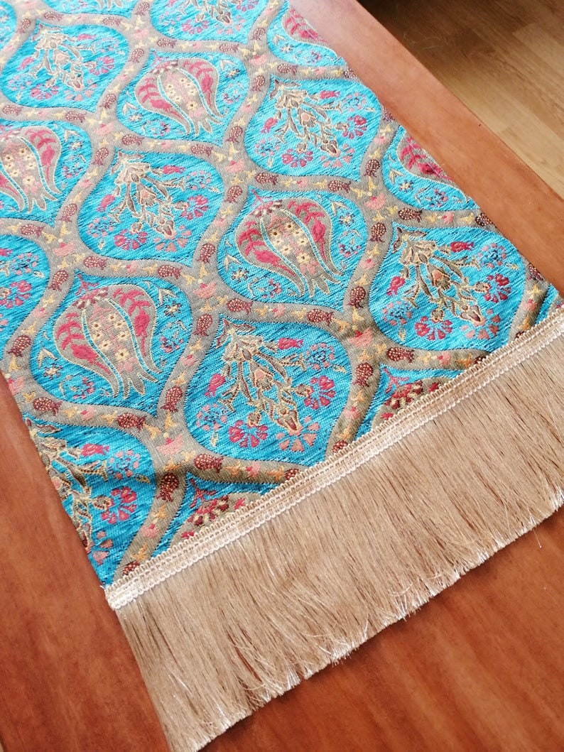 May include: A turquoise table runner with a paisley pattern and gold accents. The runner has a fringe border.