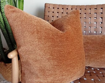 May include: Two rust-colored throw pillows with a textured fabric sit on a woven leather chair. The pillows are square and plush, offering a cozy aesthetic. A plant in a woven basket is visible in the background, adding a natural element.