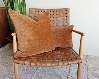 May include: A woven leather chair with two orange-brown velvet pillows. The chair has a light wood frame and a woven back and seat. The pillows are rectangular and square, adding a touch of warmth and texture to the neutral-toned room.