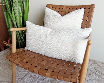 May include: Two white textured throw pillows rest on a brown leather woven chair. The chair has wooden legs and armrests. A tall green plant is visible in the background, adding a touch of nature to the scene. The pillows have a soft, inviting appearance.