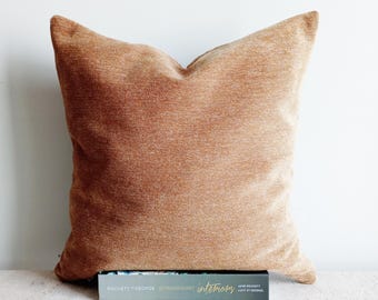 May include: A large, square, textured pillow in a warm, earthy orange-brown hue. The pillow is placed on a light-colored surface. The pillow is resting on a book with the text "ROCHETT Y GEORGE EXTRAORDINARY interiors".