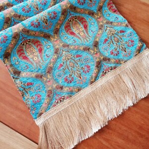 May include: A turquoise table runner with a floral pattern and gold fringe. The runner has a repeating pattern of stylized flowers and leaves.