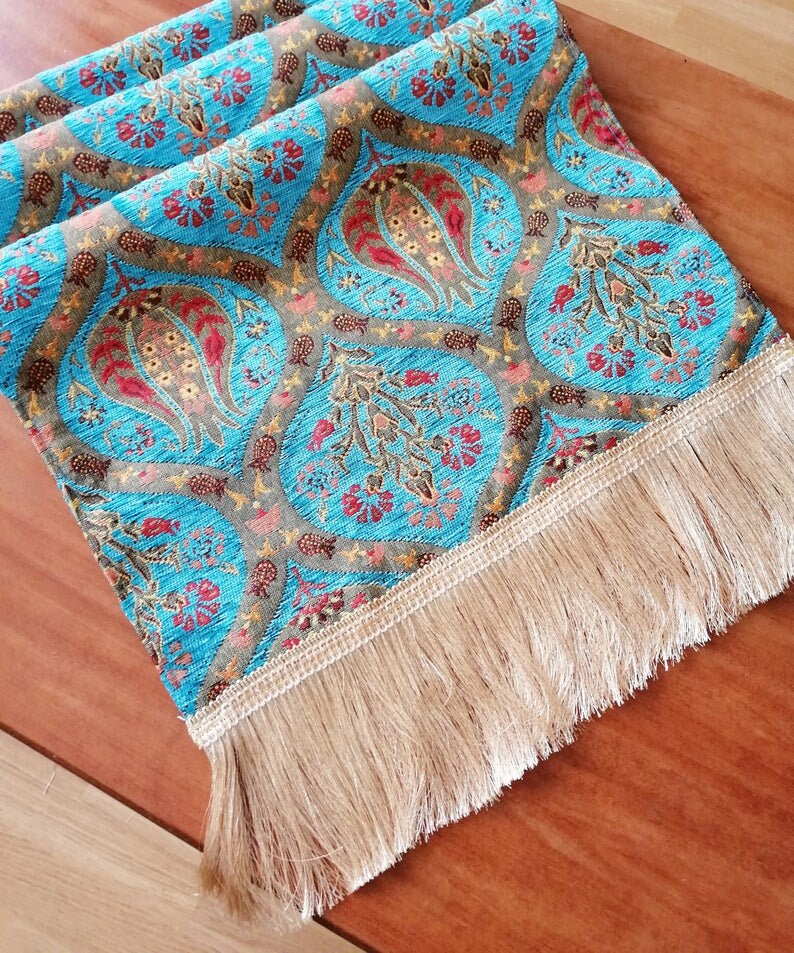 May include: A turquoise table runner with a floral pattern and gold fringe. The runner has a repeating pattern of stylized flowers and leaves.