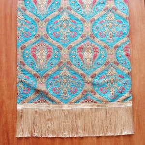 May include: A turquoise table runner with a traditional floral pattern in shades of red, gold, and brown. The runner has a fringe border.
