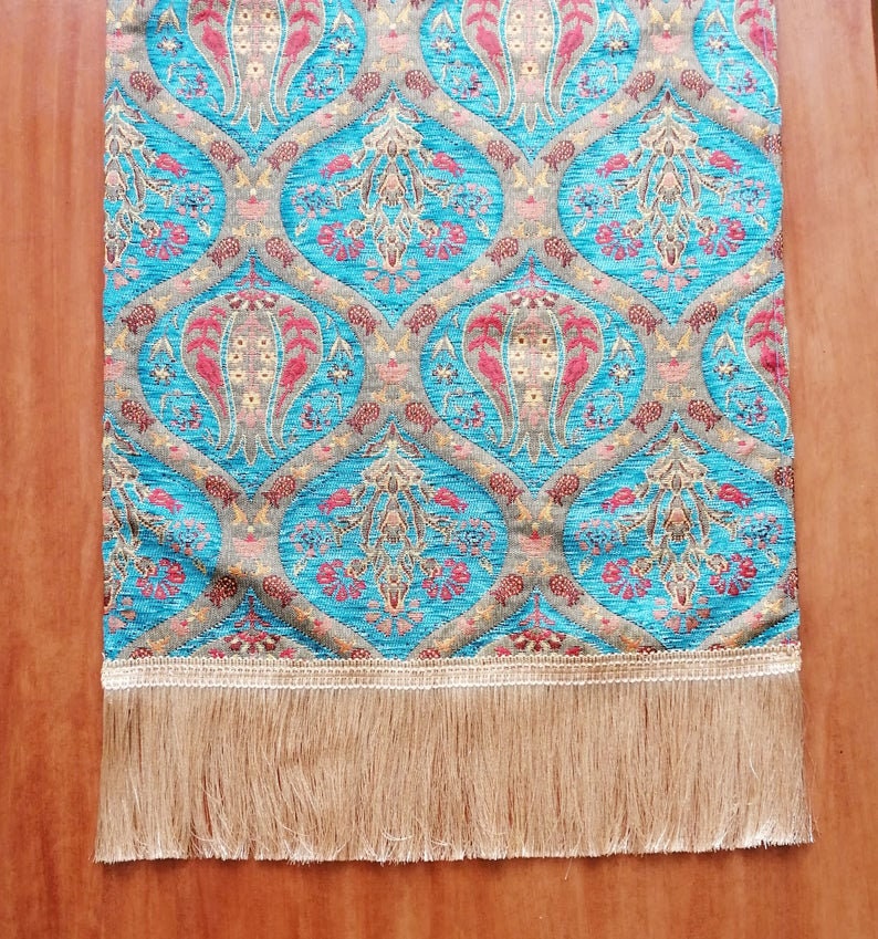 May include: A turquoise table runner with a traditional floral pattern in shades of red, gold, and brown. The runner has a fringe border.