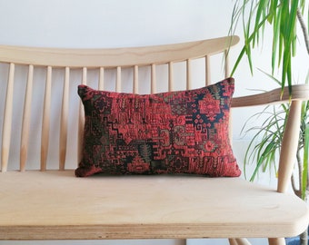 May include: Rectangular decorative pillow with a red, black, and olive green geometric pattern. The pillow is placed on a light-colored wooden bench. The pillow's design has a vintage or ethnic aesthetic.