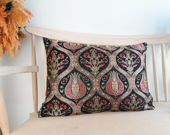 May include: Rectangular decorative pillow with a black background and a repeating floral pattern in red, gold, and gray. The pillow is placed on a light-colored wooden chair, suggesting a home decor item.