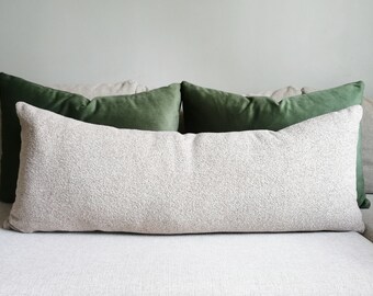 May include: A long, textured, light beige pillow is centered on a light-colored sofa, with two dark green pillows behind it. The pillows add a touch of color and texture to the neutral-toned setting.