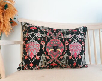May include: A black pillow with a red, gold, and silver floral pattern. The pillow is on a light wood bench.