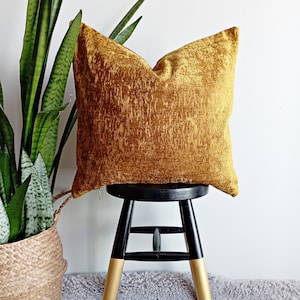 May include: A mustard yellow velvet pillow sits on a black and gold stool. The stool is on a gray rug. A green plant is in a woven basket to the left of the stool.