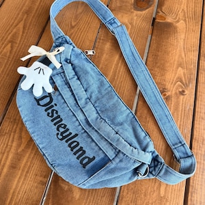 May include: A denim fanny pack with the word "Disneyland" printed in black. The bag has a matching denim strap and a white glove charm with a cream-coloured bow. The bag is on a wooden surface.