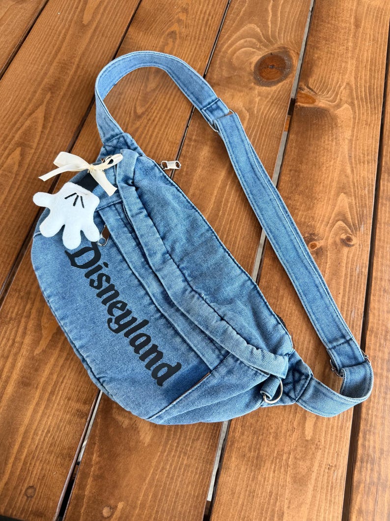May include: A denim fanny pack with the word "Disneyland" printed in black. The bag has a matching denim strap and a white glove charm with a cream-coloured bow. The bag is on a wooden surface.