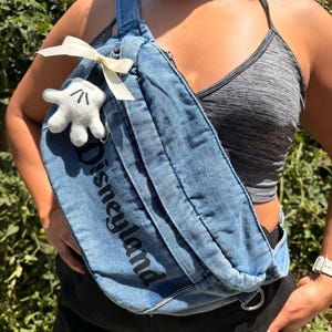 May include: A blue denim fanny pack with the word "Disneyland" printed in black. The bag features a white bow and a small white Mickey Mouse glove. The bag is worn across the body.