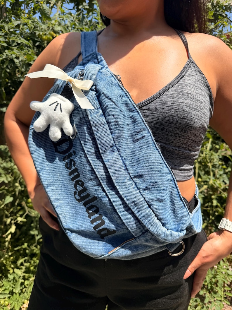 May include: A blue denim fanny pack with the word "Disneyland" printed in black. The bag features a white bow and a small white Mickey Mouse glove. The bag is worn across the body.