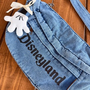May include: A blue denim fanny pack with the word "Disneyland" printed in black. A white Mickey Mouse glove-shaped charm with a cream-coloured bow is attached to the zipper. The bag has a zip closure and an adjustable strap.