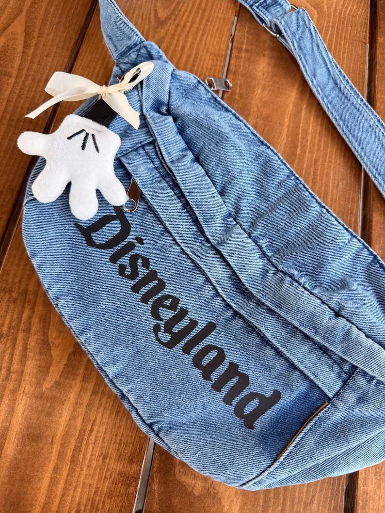May include: A blue denim fanny pack with the word "Disneyland" printed in black. A white Mickey Mouse glove-shaped charm with a cream-coloured bow is attached to the zipper. The bag has a zip closure and an adjustable strap.