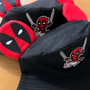 May include: Black bucket hats with a red Deadpool mask design and a cartoon Deadpool graphic with swords. The hats have a red brim and a black body, perfect for fans of the comic book character.