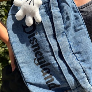 May include: A blue denim fanny pack with the word "Disneyland" printed in black. A white plush Mickey Mouse glove charm is attached to the strap. The bag is worn across the body.