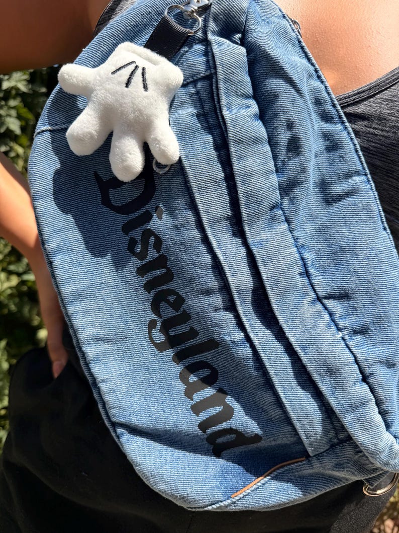 May include: A blue denim fanny pack with the word "Disneyland" printed in black. A white plush Mickey Mouse glove charm is attached to the strap. The bag is worn across the body.