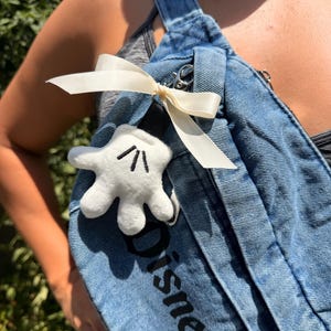 May include: A light blue denim overall strap with the word "Disneyland" printed in black. A white Mickey Mouse glove plushie is attached with a cream-coloured ribbon. The overall strap is a light blue wash.