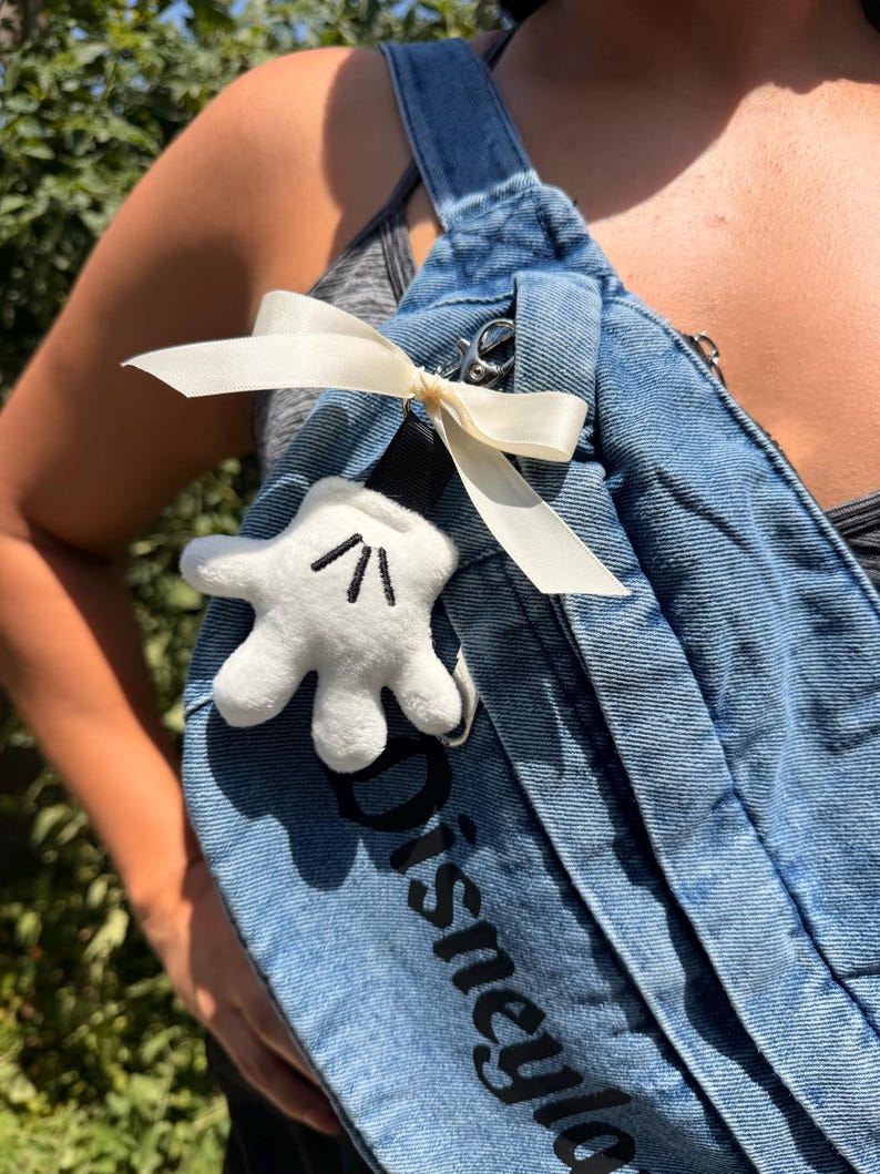 May include: A light blue denim overall strap with the word "Disneyland" printed in black. A white Mickey Mouse glove plushie is attached with a cream-coloured ribbon. The overall strap is a light blue wash.