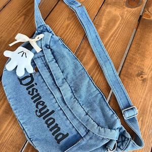May include: A blue denim fanny pack with the word "Disneyland" printed in black. The bag has a long adjustable strap and a small white Mickey Mouse glove charm with a cream-colored bow. The bag is on a wooden surface.