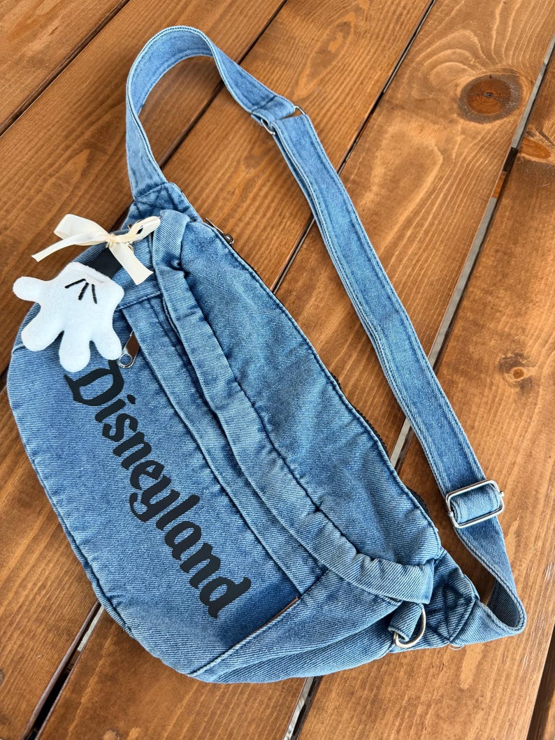 May include: A blue denim fanny pack with the word "Disneyland" printed in black. The bag has a long adjustable strap and a small white Mickey Mouse glove charm with a cream-colored bow. The bag is on a wooden surface.