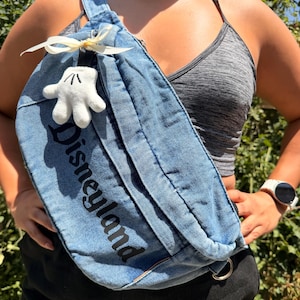 May include: A blue denim fanny pack with the word "Disneyland" printed in black. The bag features a white Mickey Mouse glove charm and a cream-coloured bow. The bag is worn across the body.