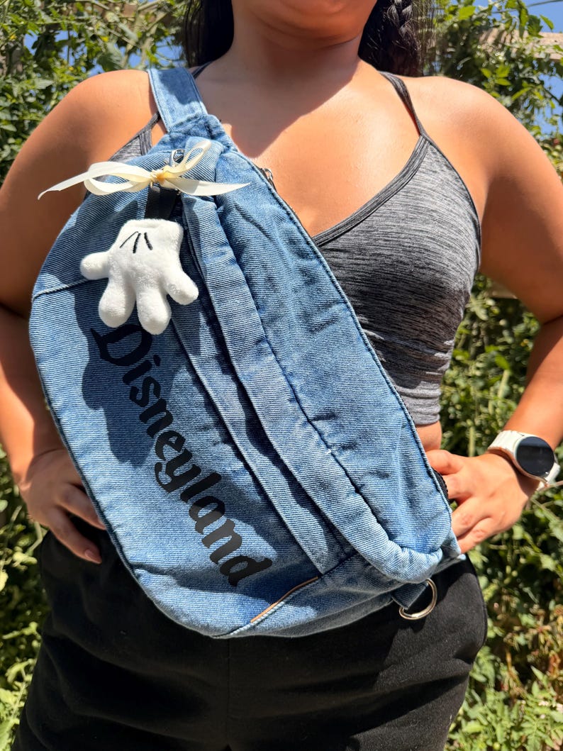 May include: A blue denim fanny pack with the word "Disneyland" printed in black. The bag features a white Mickey Mouse glove charm and a cream-coloured bow. The bag is worn across the body.