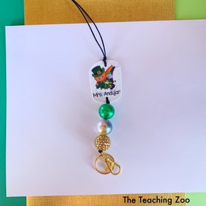 May include: A green and gold lanyard with a charm featuring a cartoon leprechaun and the text "Mrs. Andujar". The lanyard has a gold clasp and three beads: one green, one iridescent, and one gold.
