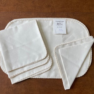 May include: A set of off-white linen placemats and napkins with a decorative trim. The set includes a large oval placemat, three square placemats, and three folded napkins. A tag reads "Permanent Press" and includes fabric details.
