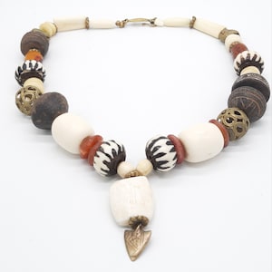 May include: A white and brown beaded necklace with a bone pendant and a gold arrow charm. The necklace features a variety of beads, including round, cylindrical, and carved beads.