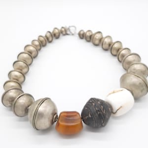 May include: A silver beaded necklace with a silver clasp. The necklace has three accent beads: one amber, one black with a carved design, and one white.
