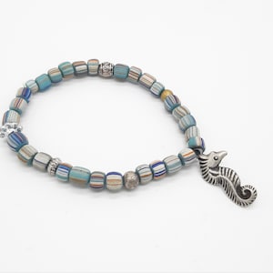 Puede incluir: Un brazalete de cuentas azules y blancas con un dije de caballito de mar plateado. Las cuentas son multicolores y tienen un patrón a rayas.