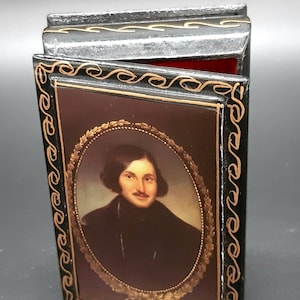 Op de afbeelding: Een zwarte houten doos met een gouden rand en een portret van een man erin. De doos is versierd met een gouden patroon.